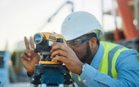 On a commercial construction site, a professional engineer surveyor takes measures with theodolite in the blurry background are steel formwork frames and a crane.