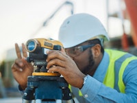 On a commercial construction site, a professional engineer surveyor takes measures with theodolite in the blurry background are steel formwork frames and a crane.