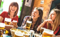 Group of women laughing and joking in a pub with several full pints of lager and food on the table