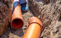 Close up of laying drainage pipes into the ground. Drainpipes are orange and sit in a soil trench.