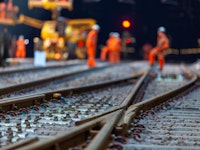 Low angle shot of some cross roads on a railway station tracks with workers wearing orange hi-vis vests blurred in the background.