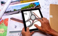 Town and land use planning. An employee holding in his hands a digital tablet displaying a road project plan, above a desk where ground plan maps are placed.