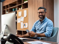 Smiling business man wearing eyeglasses working on desktop computer while using phone in office