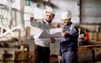 Two male engineers are standing in the building site and holding construction plan. Businessmen dressed in white protective hardhats working together and discussing details of a design project.